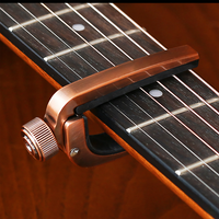A close-up of a metallic bronze guitar capo clamped onto a dark wood guitar neck with strings visible.