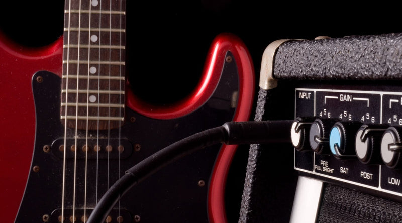 Against a black background, this close-up shows a glossy red electric guitar connected via an audio cable to a guitar amp fitted with multiple tone control knobs.