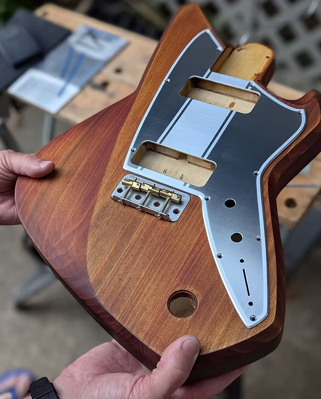 Wooden guitar body with metal hardware held by a person on a blurred workshop background