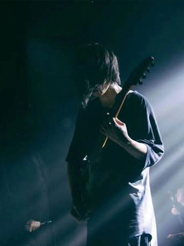 A person in a dark oversized shirt holding an electric guitar on a dim stage with dramatic spotlighting and haze.