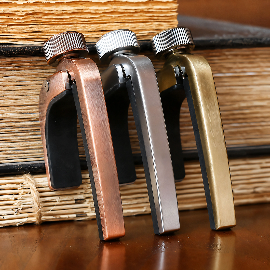 Three metal bookends with screw-top handles in copper, silver, and gold finishes, arranged side by side on a wooden surface in front of stacked books.