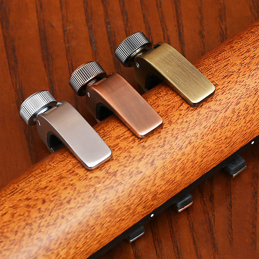 Three metal guitar tailpiece hardware pieces with knurled adjustment knobs, shown mounted on a wooden guitar headstock against a dark wood background.