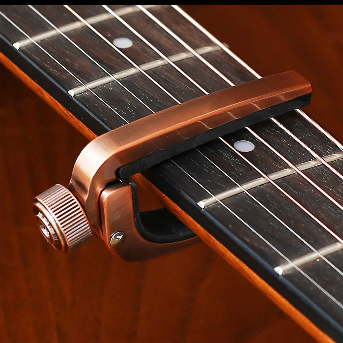 A close-up of a metallic bronze guitar capo clamped onto a dark wood guitar neck with strings visible.