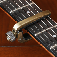 A close-up of a brass guitar capo clamped onto the neck of an acoustic guitar, with the adjustment knob visible on the left.