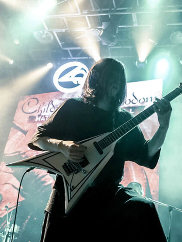 A musician with long dark hair plays a white electric guitar on stage under dramatic stage lighting and smoke. A large circular band logo is visible in the background above the performer.